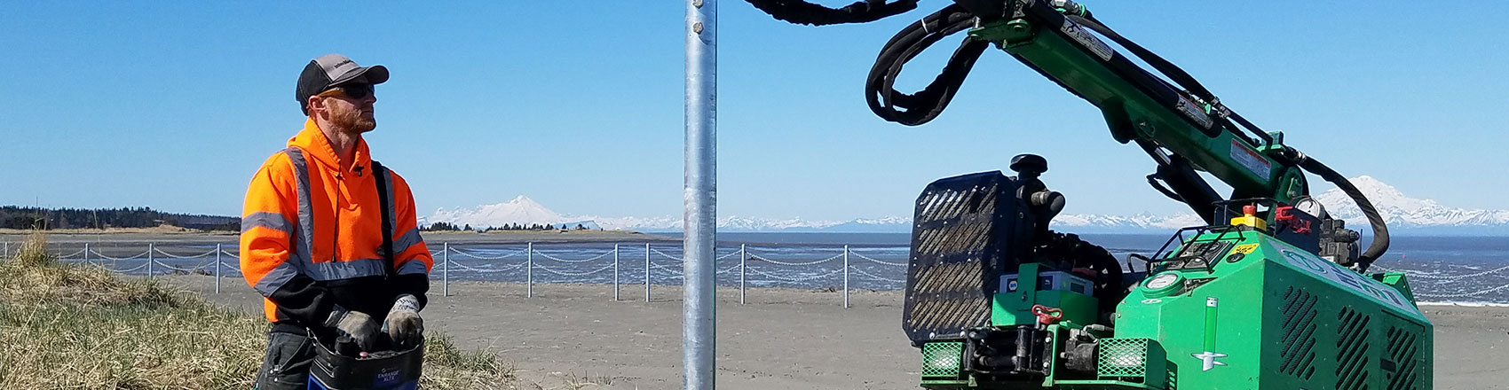 Crew member installing metal post on Alaskan coastline with volcanoes in the background.